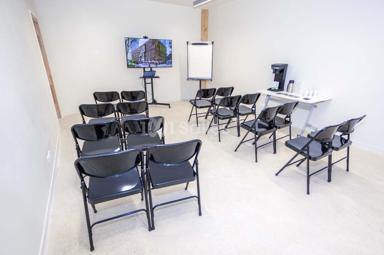 Salle de réunion avec chaises noires alignées, écran, paperboard et table équipée d'une machine à café et gobelets.