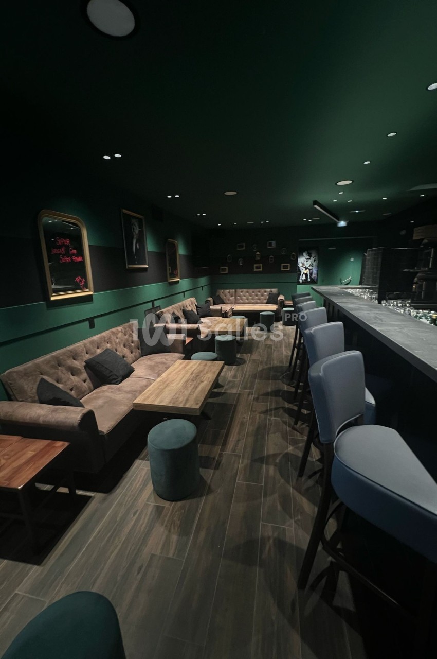 Intérieur d'un bar avec des fauteuils, tables basses, tabourets hauts et un éclairage tamisé dans une ambiance sombre.