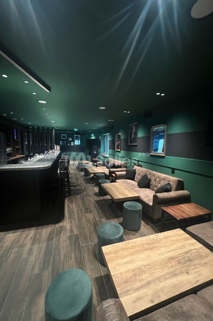 Salle lounge moderne avec canapés, tables basses en bois, tabourets et un bar éclairé dans une ambiance tamisée.