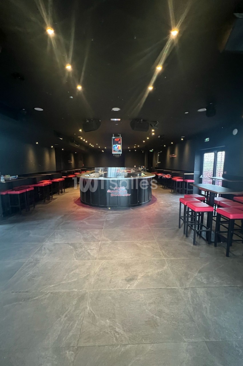 Salle sombre avec un bar central arrondi, entouré de tables hautes et de tabourets rouges.
