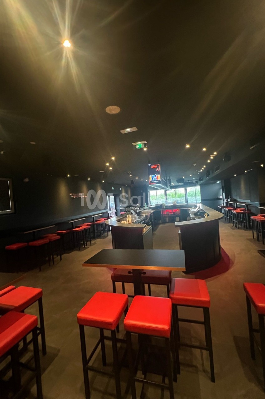 Salle de bar moderne avec tables hautes, tabourets rouges et éclairage tamisé, vue sur un comptoir central.