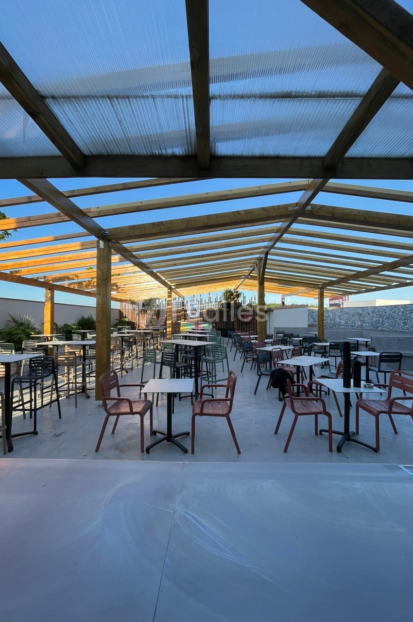 Terrasse extérieure couverte avec tables et chaises espacées, entourée de plantes et éclairée par la lumière naturelle.