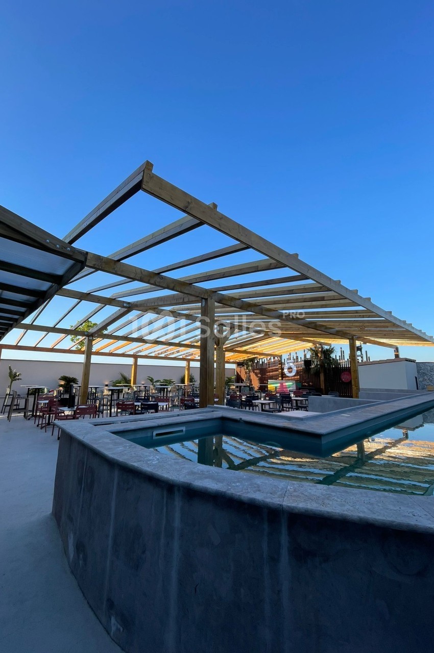 Terrasse extérieure avec piscine, pergola en bois et tables disposées pour un espace de détente sous un ciel dégagé.
