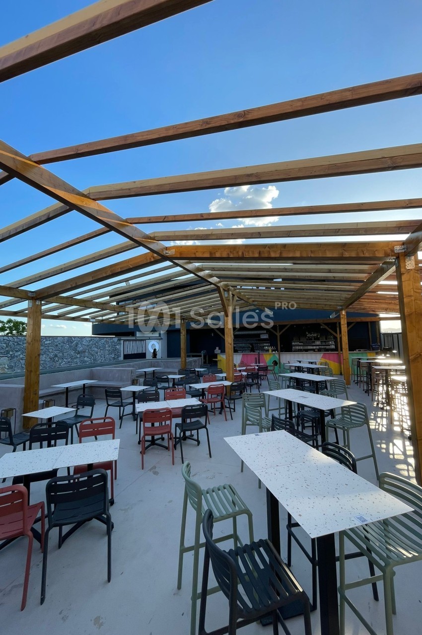 Terrasse extérieure avec tables et chaises colorées sous une structure en bois ouverte, ciel bleu en arrière-plan.