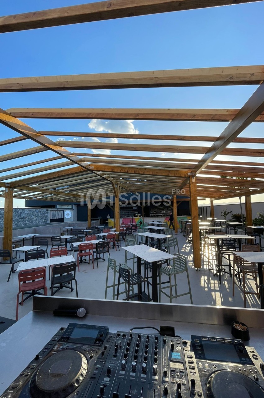 Terrasse extérieure ensoleillée avec tables, chaises et une cabine de DJ au premier plan sous une structure en bois.