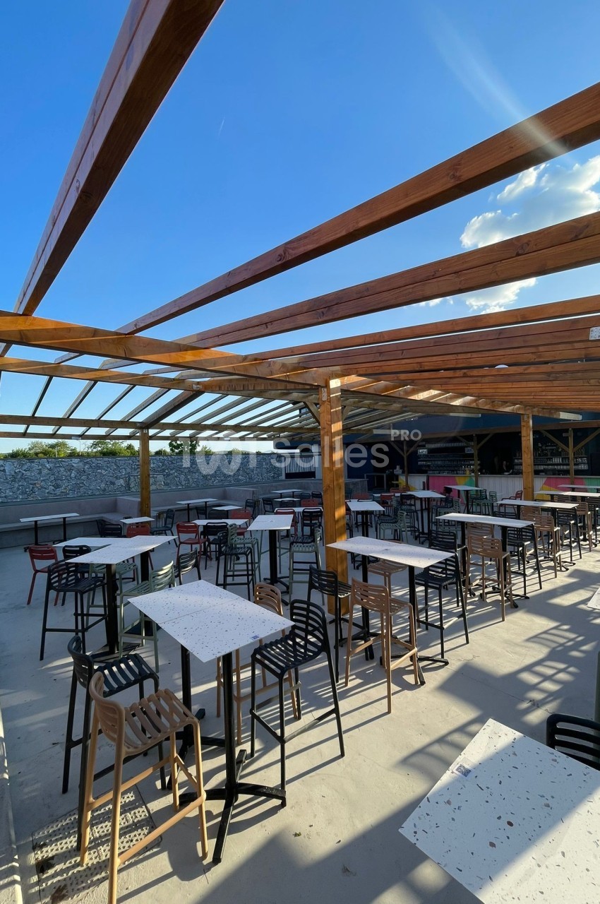 Terrasse extérieure avec tables et chaises sous une structure en bois ouverte, éclairée par un ciel ensoleillé.
