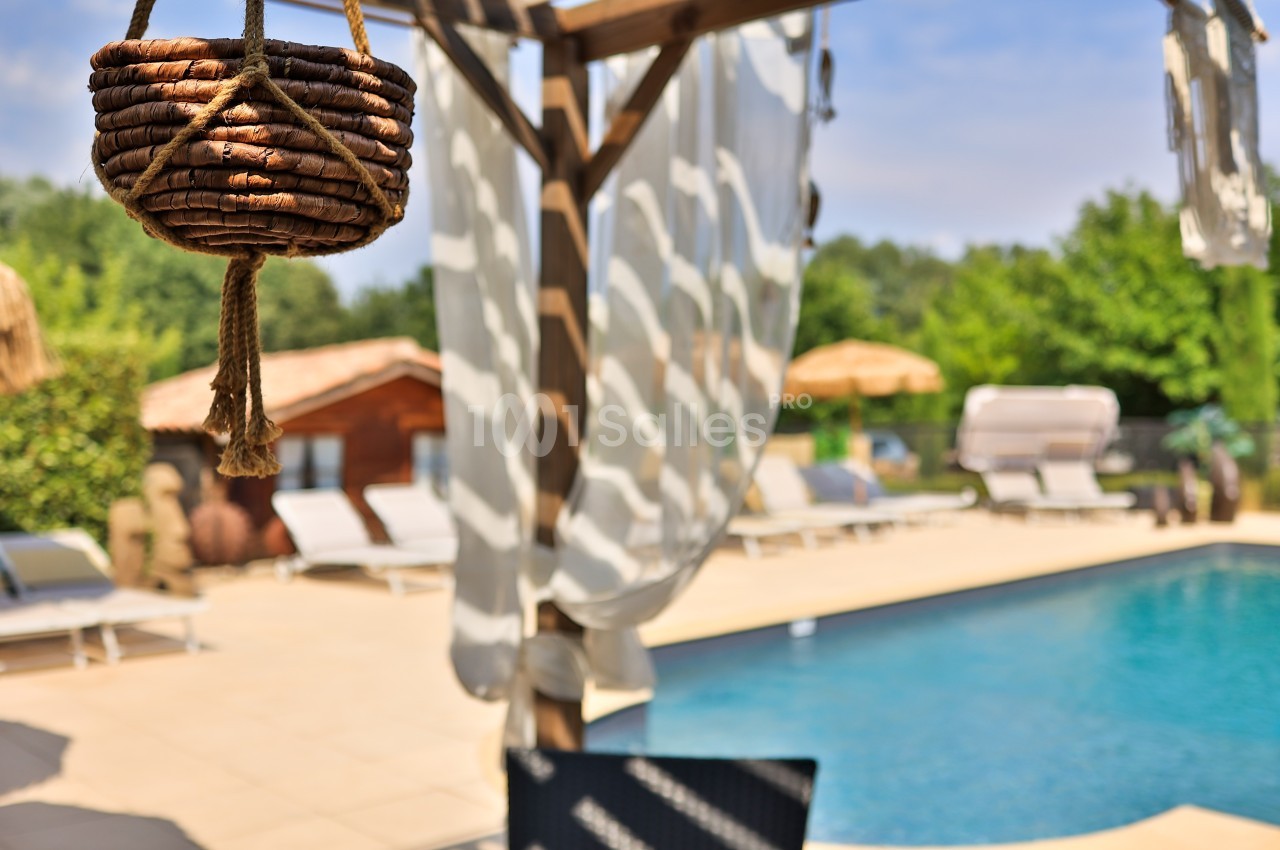 Panier suspendu en osier au premier plan, avec une piscine et des transats floutés en arrière-plan sous un ciel dégagé.