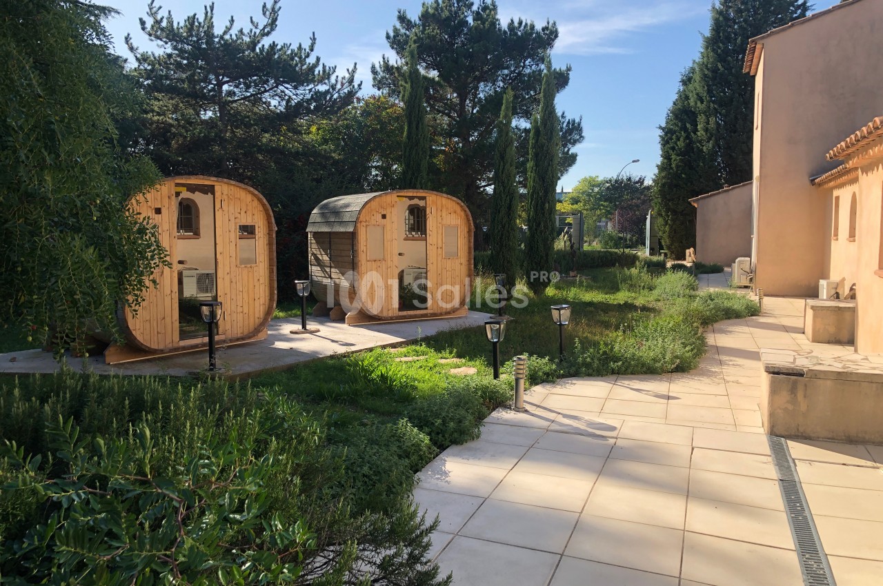 Deux cabanes en bois arrondies dans un jardin verdoyant, près d'une allée pavée et d'un bâtiment beige.