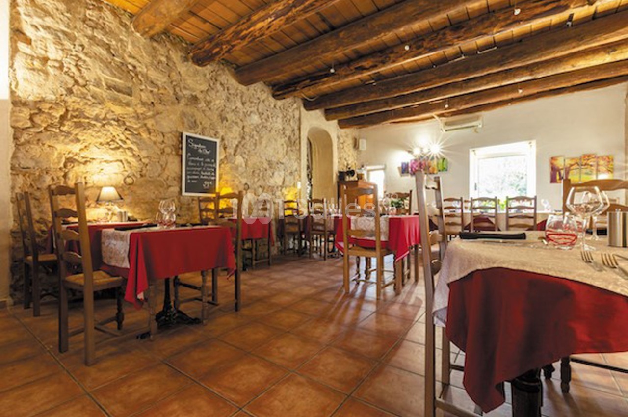 Salle de restaurant rustique avec murs en pierre, plafond en bois, tables dressées et nappes rouges.