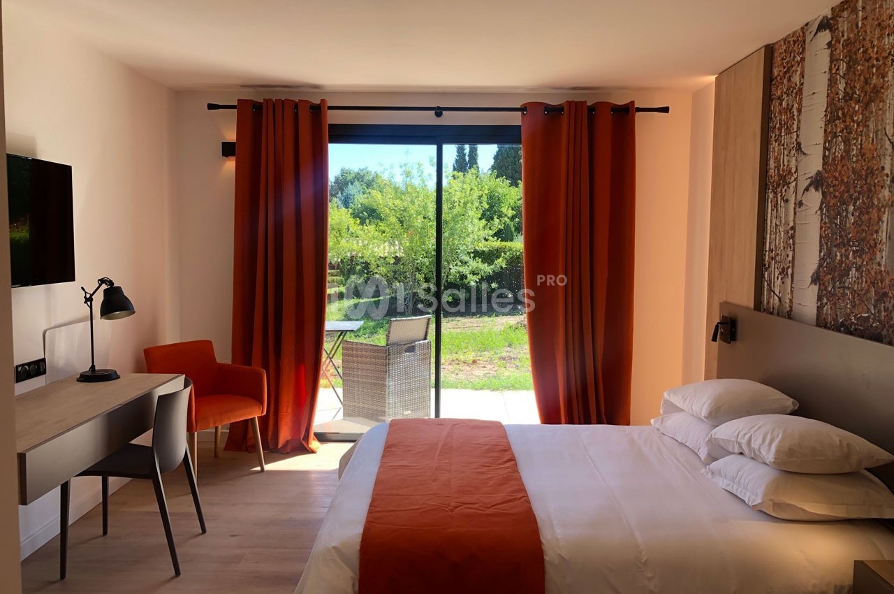 Chambre lumineuse avec lit double, bureau, chaise, rideaux orange et baie vitrée donnant sur un jardin verdoyant.