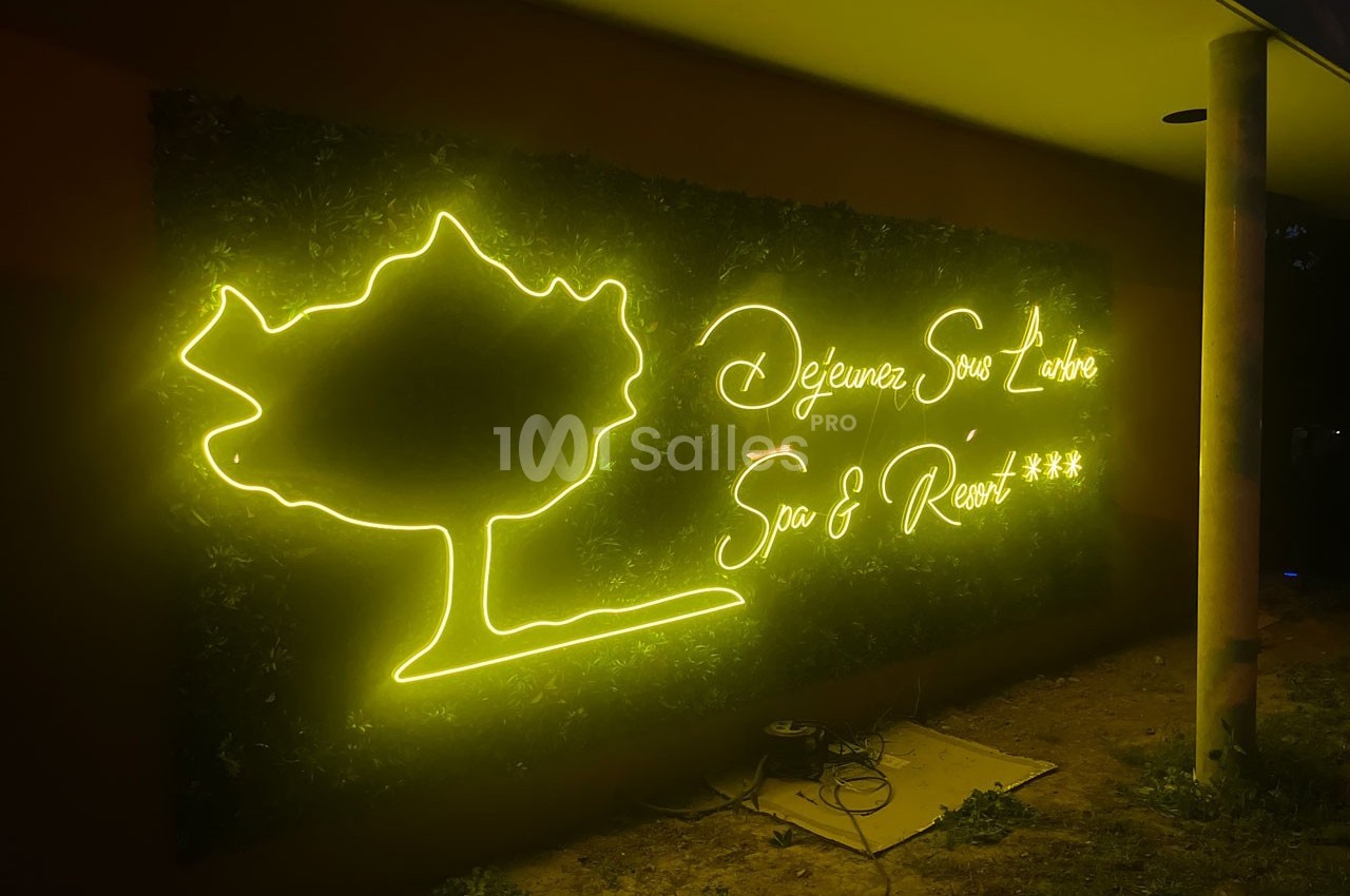 Enseigne lumineuse jaune représentant un arbre et le texte ’Déjeunez Sous l'Arbre Spa & Resort ***’ sur un mur sombre.