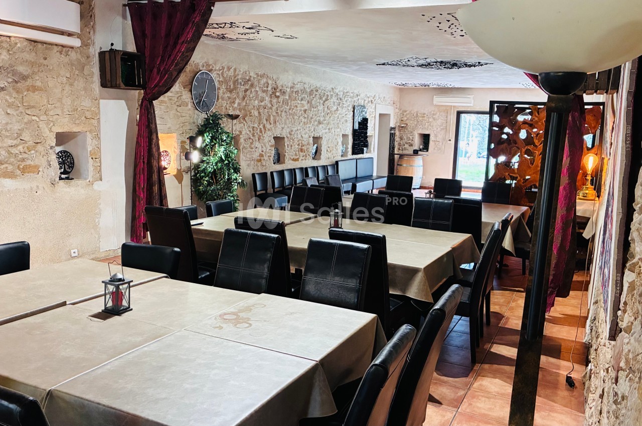 Salle de restaurant avec murs en pierre, grandes tables rectangulaires, chaises noires et lumière tamisée.