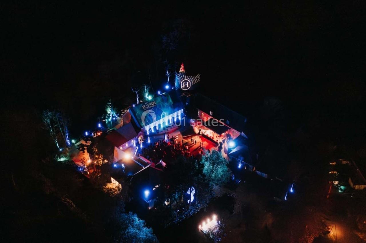 Vue aérienne nocturne d'un bâtiment éclairé par des lumières colorées entouré de végétation sombre.