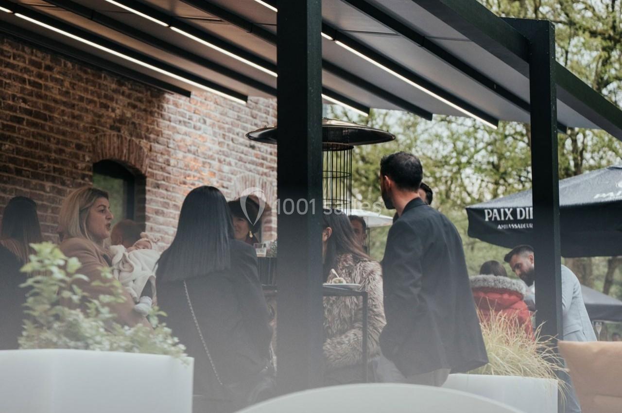 Groupe de personnes discutant sous une terrasse couverte près d'un mur en briques, entourées de plantes.