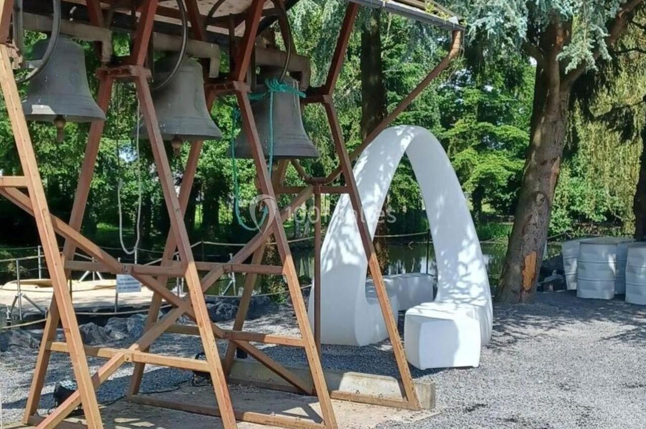 Structure en bois avec des cloches suspendues, près d'une arche blanche et d'arbres dans un espace extérieur.