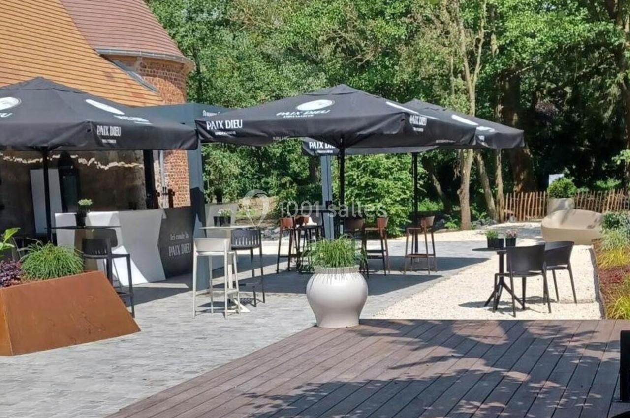 Terrasse extérieure avec tables, chaises et parasols noirs, entourée de verdure et d'un bâtiment en briques.