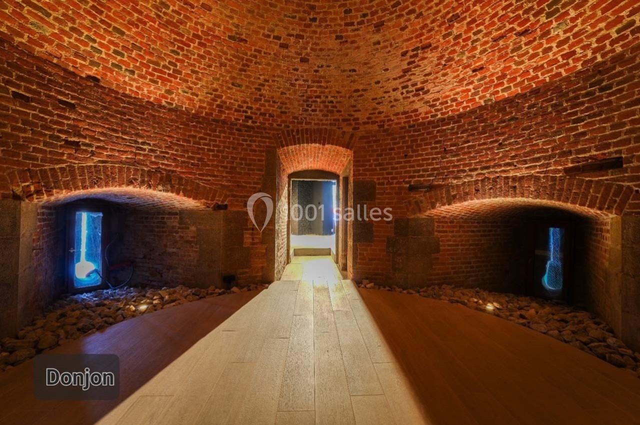 Salle voûtée en briques rouges avec éclairage tamisé, petites ouvertures et sol en bois clair.