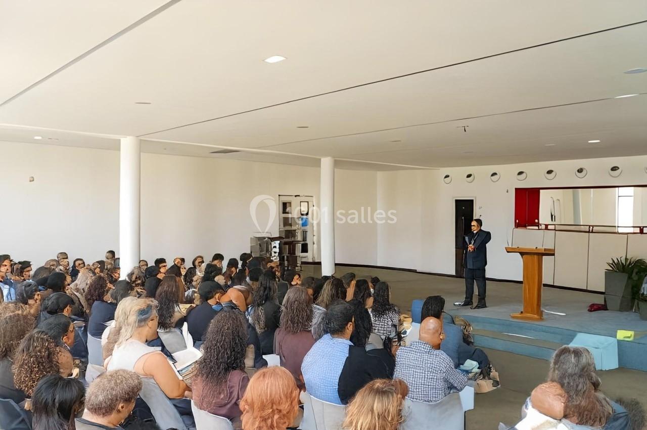 Un orateur s'adresse à un public assis dans une salle lumineuse avec un pupitre en bois au premier plan.