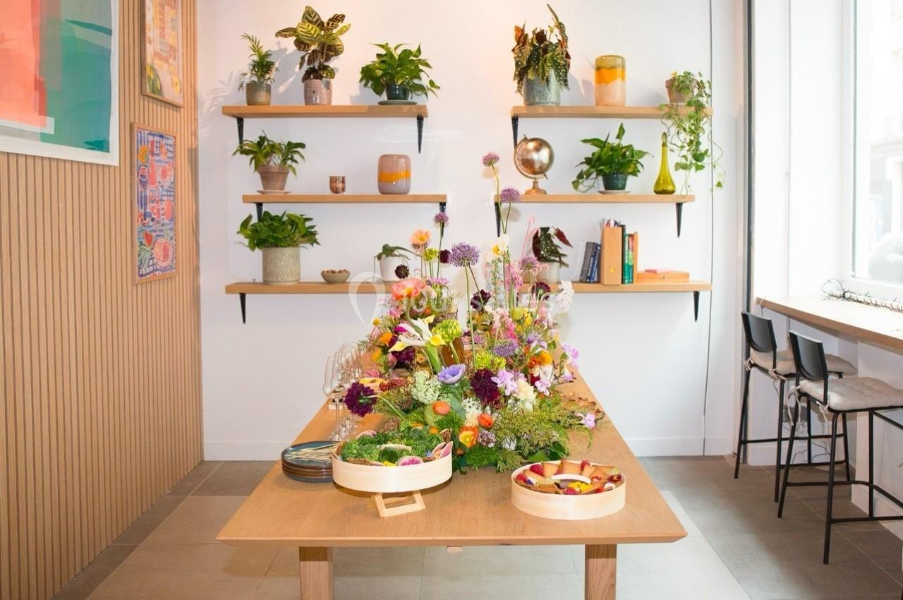 Table en bois décorée de fleurs colorées, entourée de plantes sur des étagères et de chaises près d'une fenêtre.
