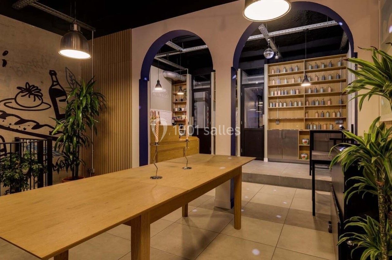 Intérieur d'un espace moderne avec une grande table en bois, des plantes et des étagères remplies de bouteilles.