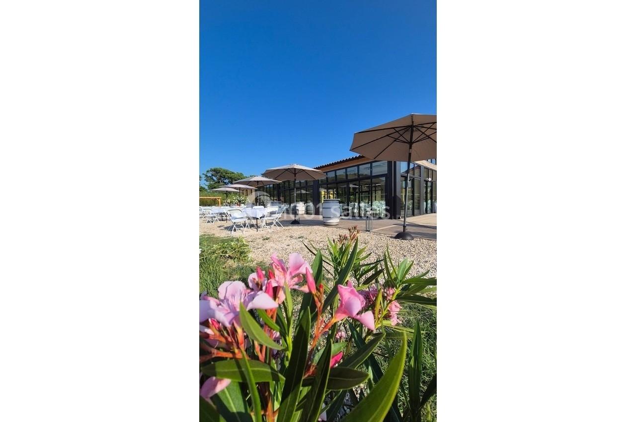 Bâtiment moderne avec terrasse ombragée, tables extérieures et fleurs roses au premier plan sous un ciel bleu.