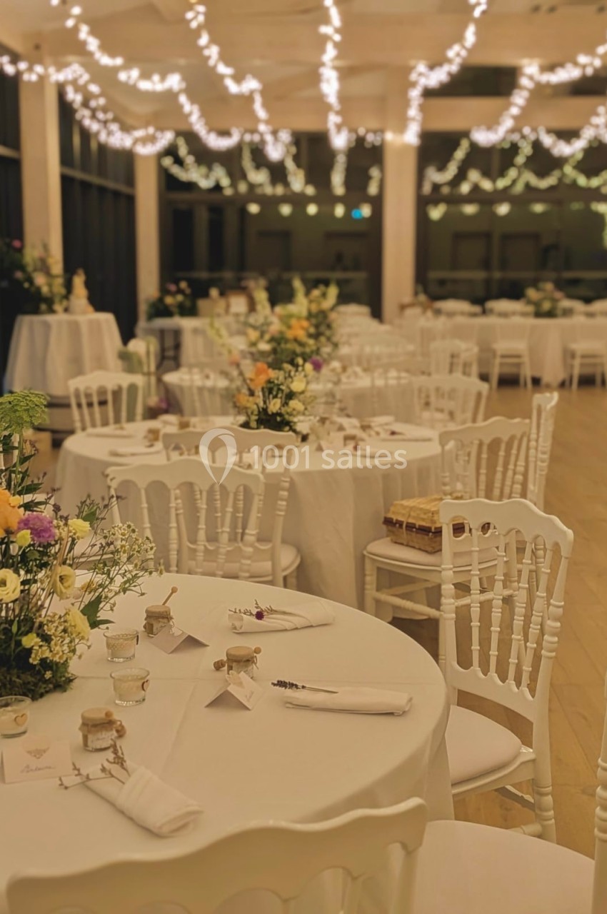 Salle de réception décorée avec des guirlandes lumineuses, tables rondes dressées et ornées de fleurs.