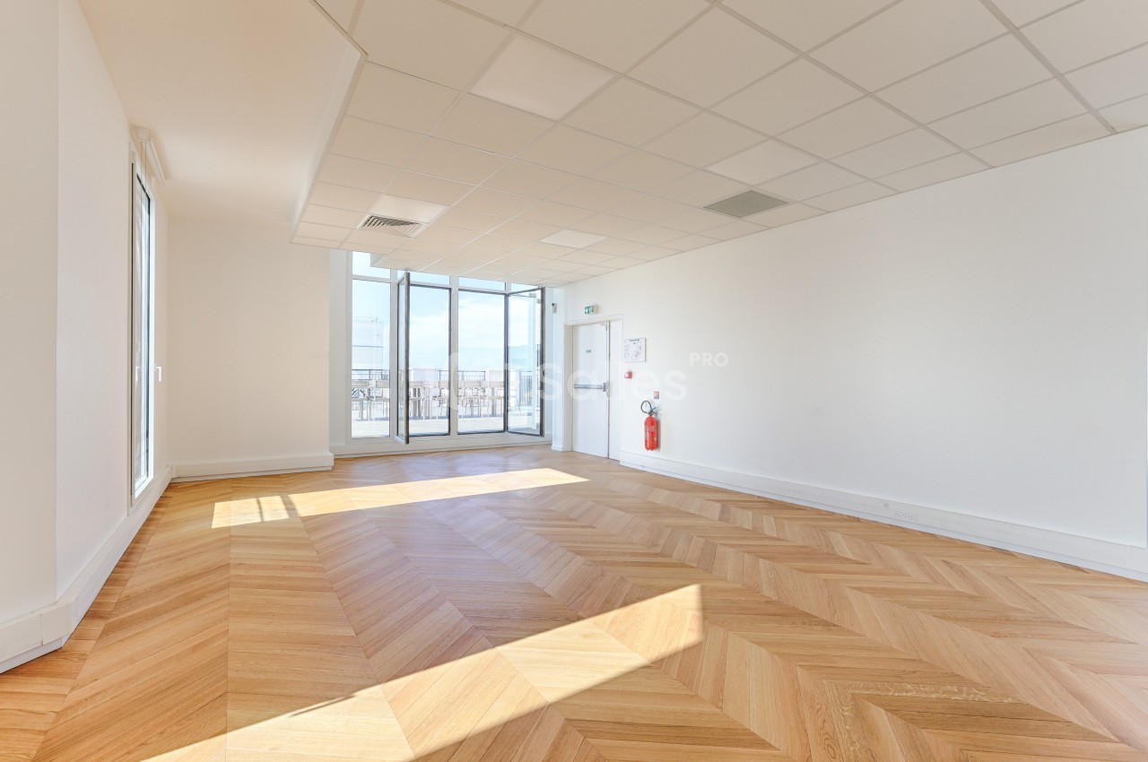 Salle lumineuse avec parquet en chevrons, grandes baies vitrées donnant sur une terrasse et plafond à dalles blanches.