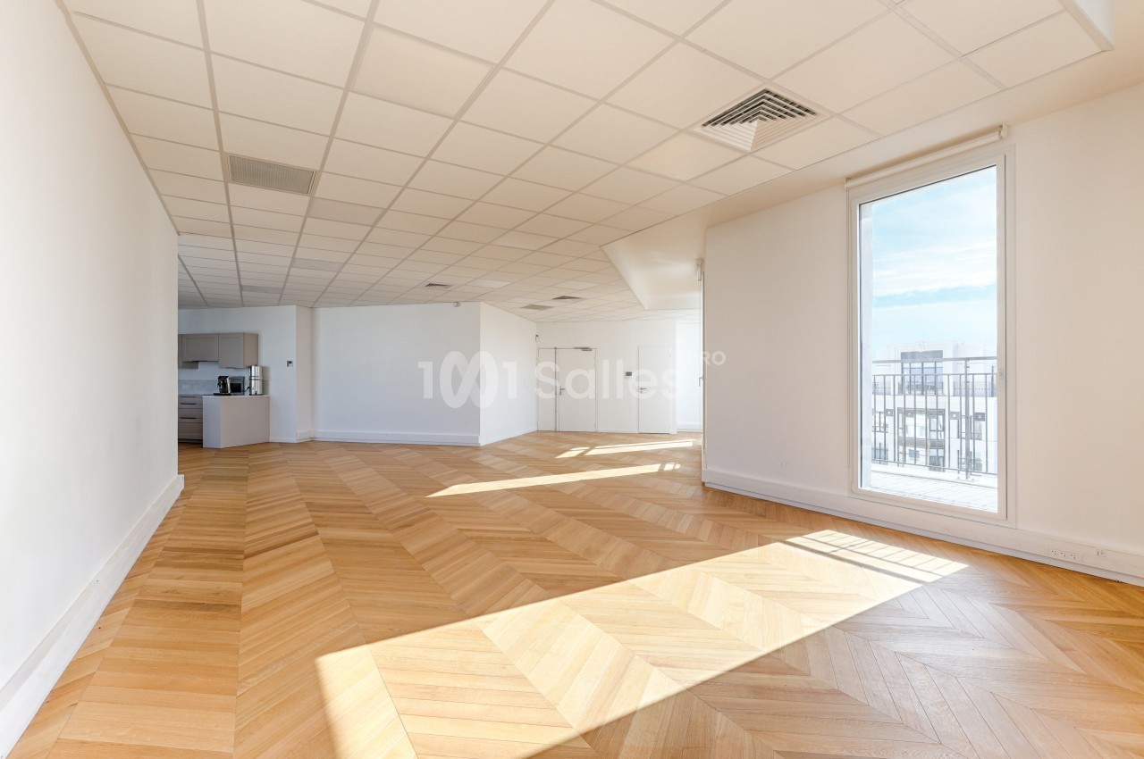 Salle lumineuse avec parquet en chevrons, plafond à dalles blanches, cuisine ouverte et grande fenêtre donnant sur un balcon.