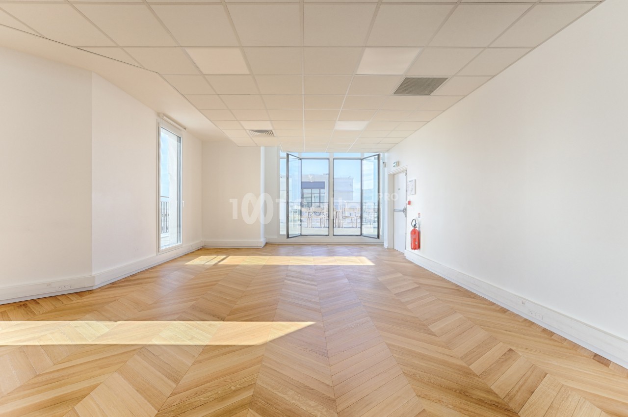 Salle lumineuse avec parquet en chevrons, murs blancs, plafond à dalles et grande baie vitrée donnant sur l'extérieur.