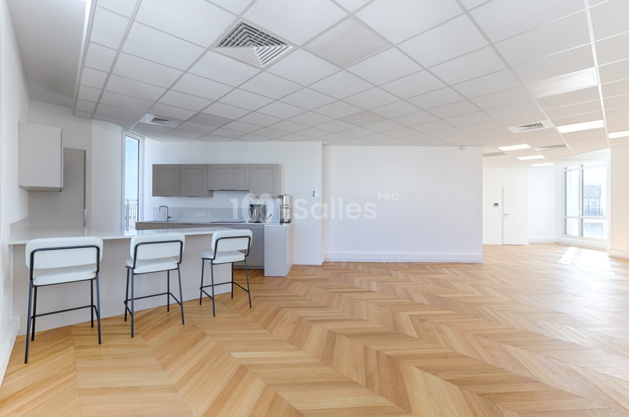 Cuisine ouverte avec comptoir et tabourets, parquet en chevrons et grande pièce lumineuse avec fenêtres.