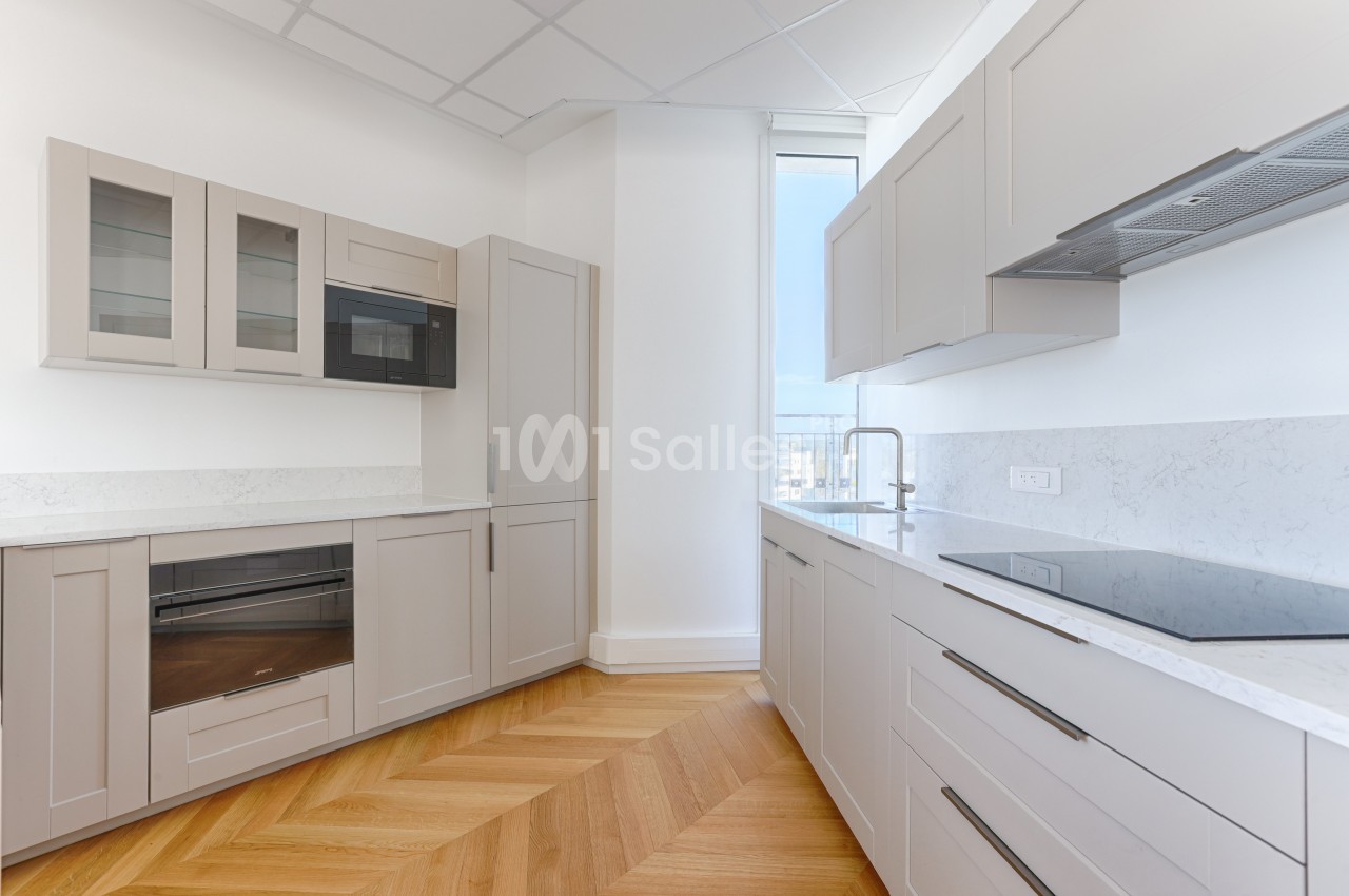 Cuisine moderne avec meubles beige clair, plan de travail en marbre blanc, parquet en bois et fenêtre lumineuse.