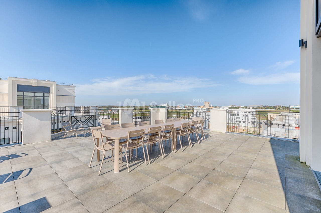 Terrasse spacieuse avec tables et chaises en bois, offrant une vue dégagée sur des bâtiments et un ciel bleu.