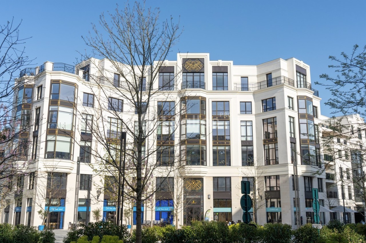 Bâtiment moderne à plusieurs étages avec des façades blanches, de grandes fenêtres et des arbres au premier plan.