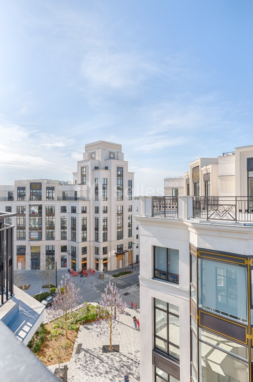 Vue sur des bâtiments modernes de style classique entourant une petite place avec des arbres en fleurs, sous un ciel dégagé.