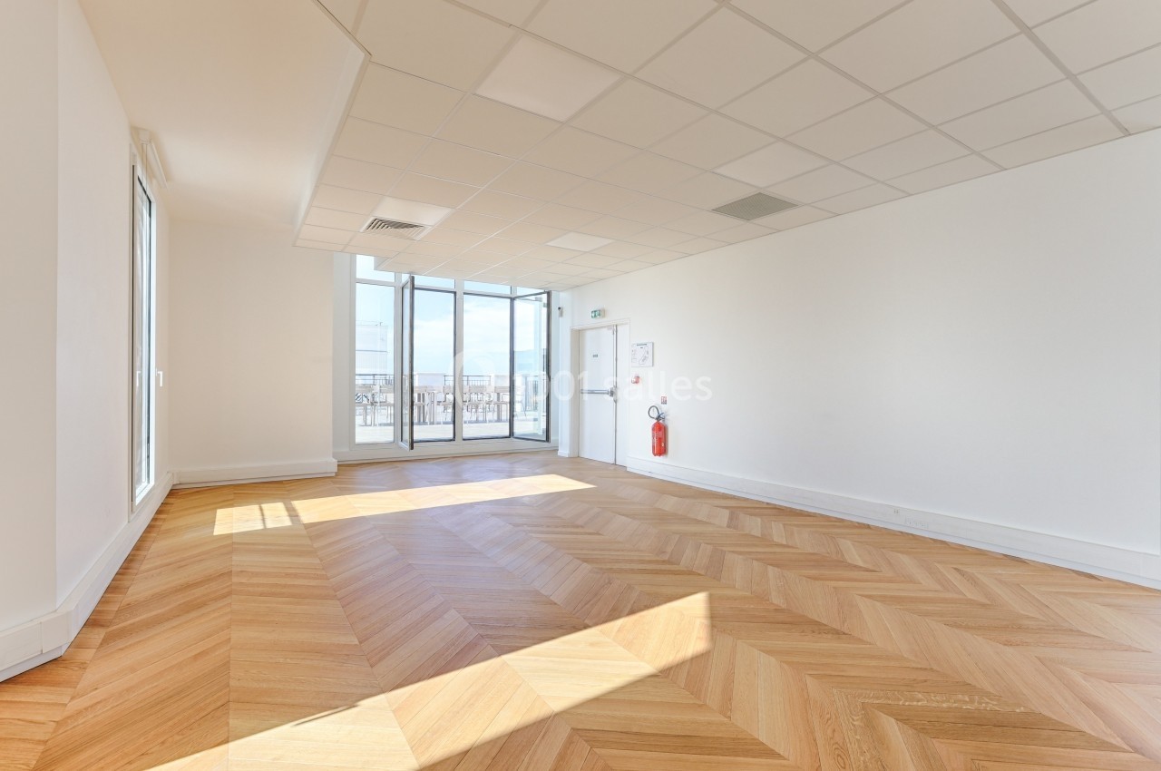Salle vide avec parquet en chevrons, murs blancs, plafond à dalles et grande baie vitrée donnant sur une terrasse.