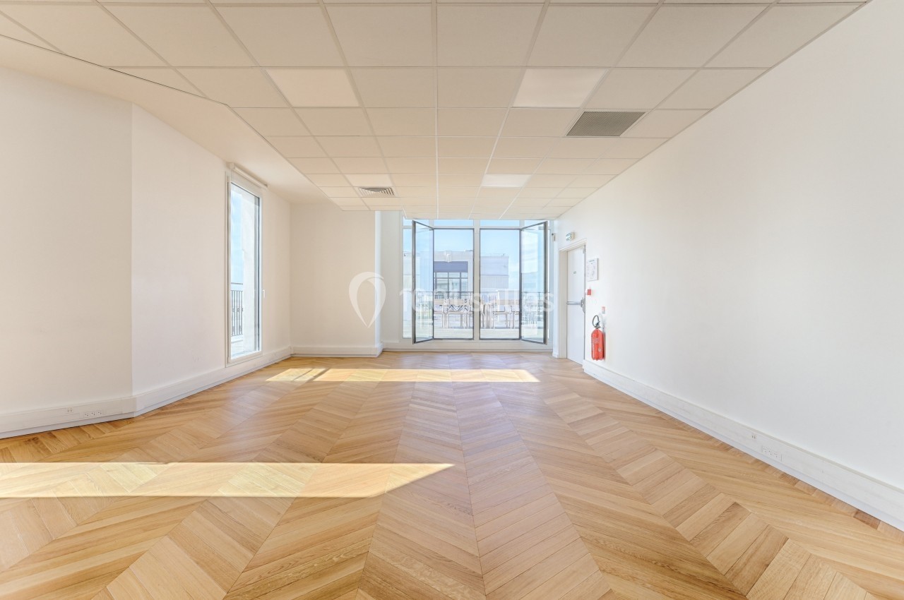 Salle lumineuse avec parquet en chevrons, murs blancs, plafond à dalles et grande baie vitrée donnant sur l'extérieur.