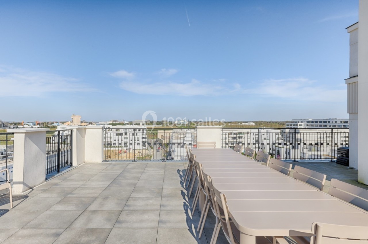 Terrasse spacieuse avec une longue table et des chaises, offrant une vue dégagée sur des bâtiments et un ciel bleu.