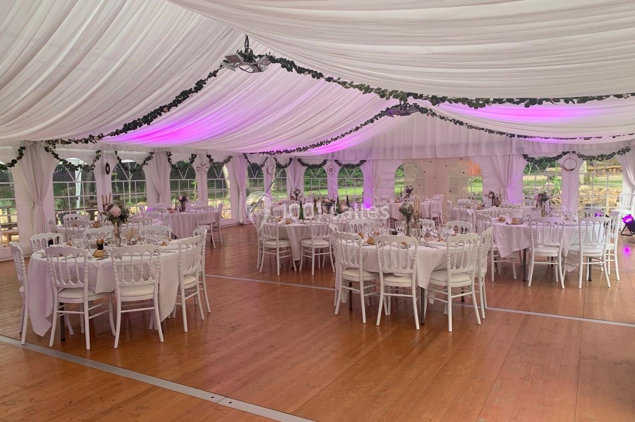 Salle de réception sous chapiteau blanc avec tables rondes décorées, chaises blanches et éclairage rose subtil.
