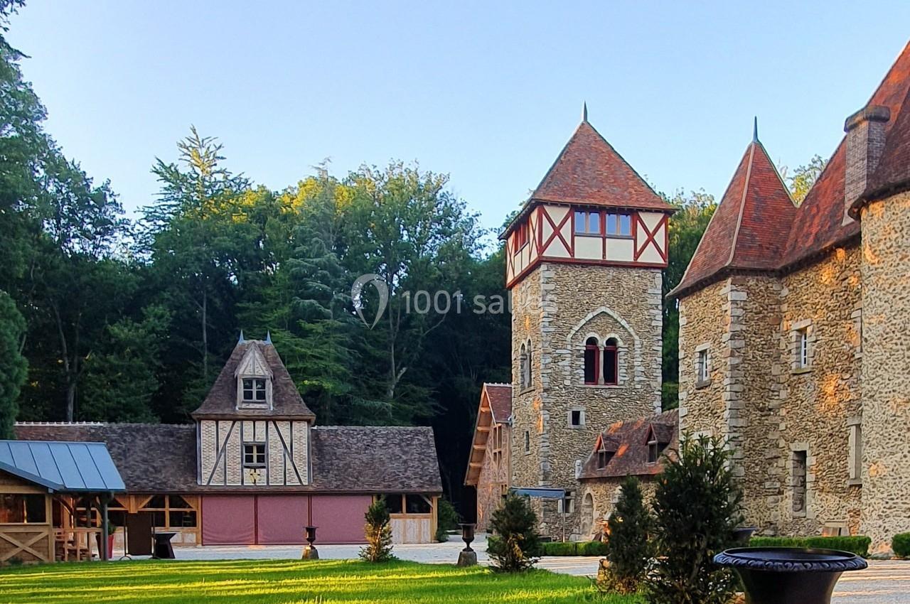 Cour d'un domaine avec bâtiments en pierre et à colombages, entourée de pelouse et bordée d'arbres sous un ciel dégagé.