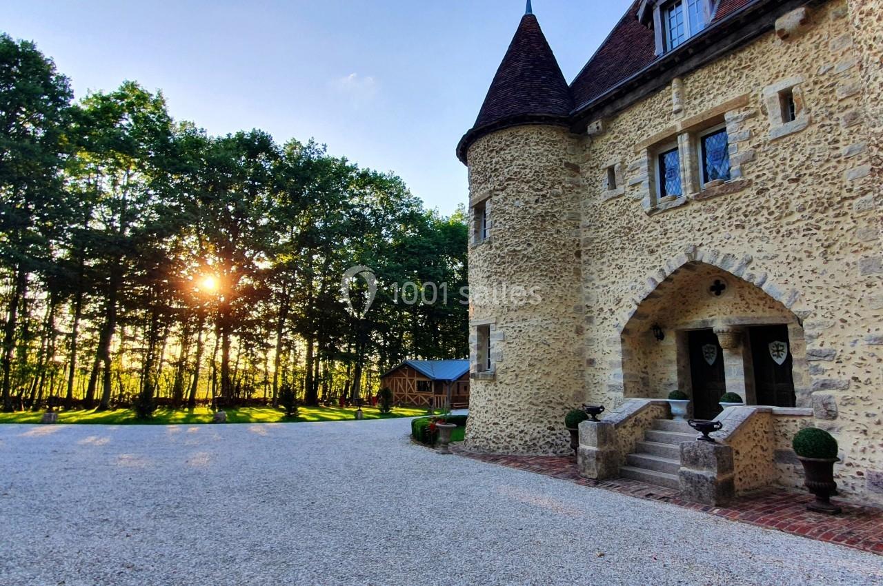Façade en pierre d'un château avec tourelles, entouré d'arbres et éclairé par un soleil couchant.
