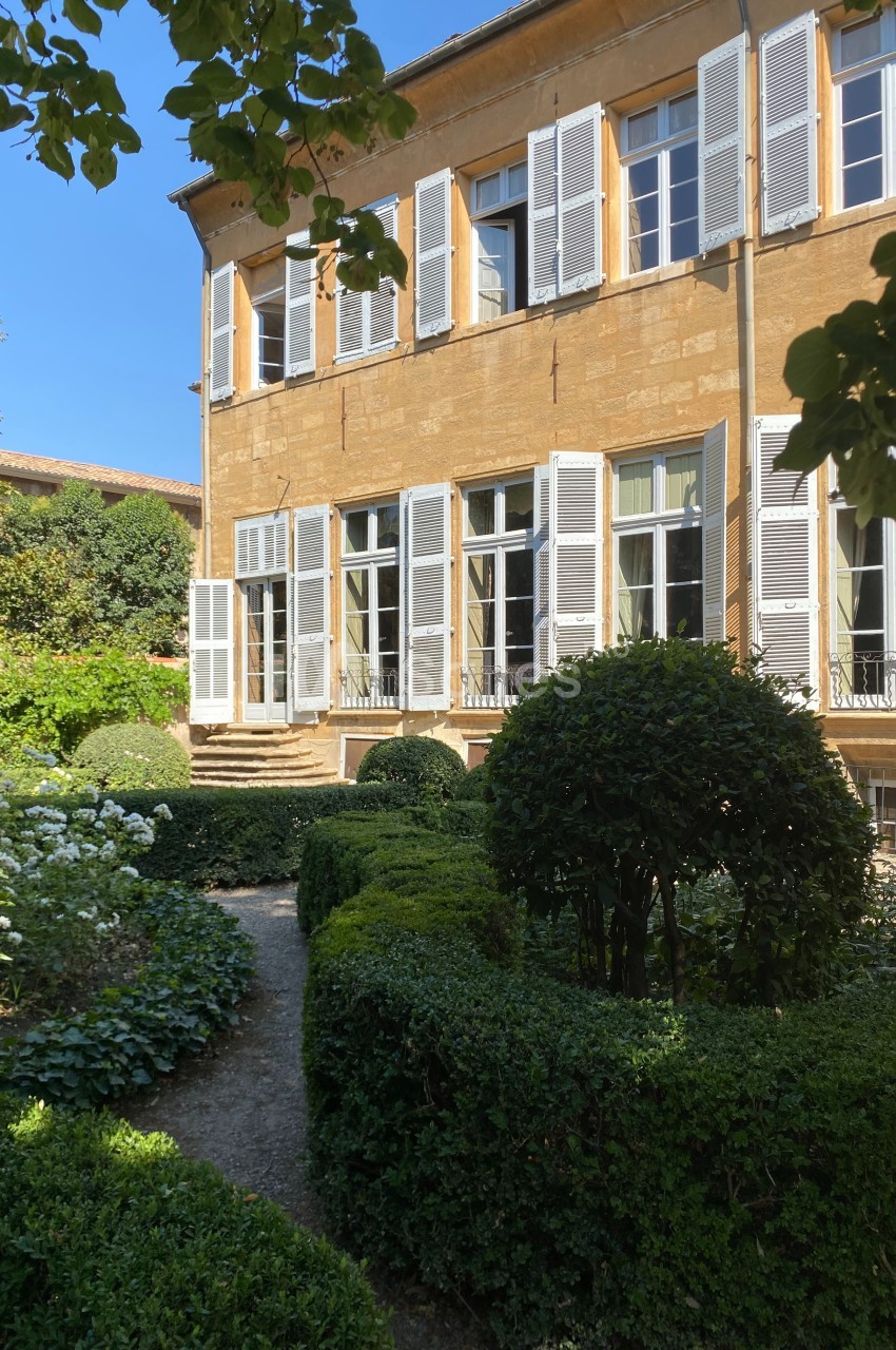 Façade d'une maison en pierre ocre avec volets blancs, entourée d'un jardin verdoyant et bien entretenu.