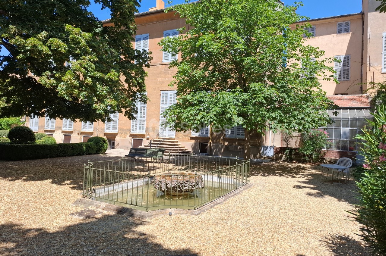 Cour avec un petit bassin entouré de graviers, bordée d'arbres et d'un bâtiment ancien aux volets blancs.