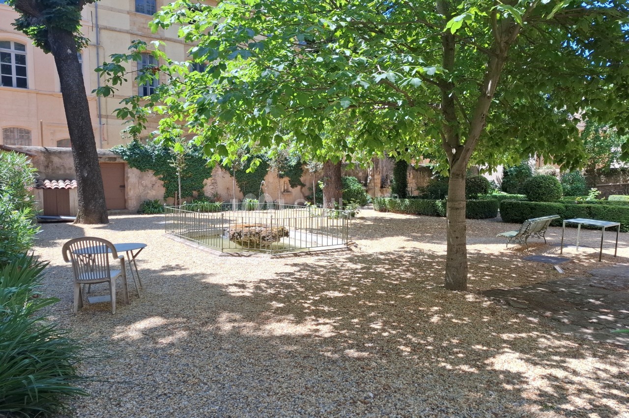 Jardin ombragé avec des arbres, des chaises en bois, une table et une petite clôture entourant un espace central.
