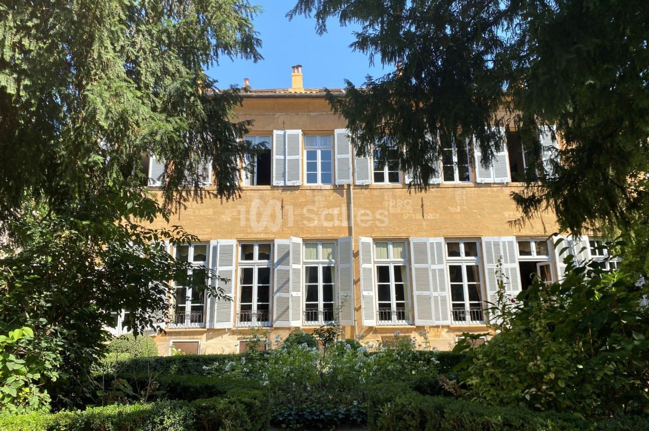 Façade d'un bâtiment ancien en pierre dorée avec volets blancs, entouré de végétation et d'arbustes.