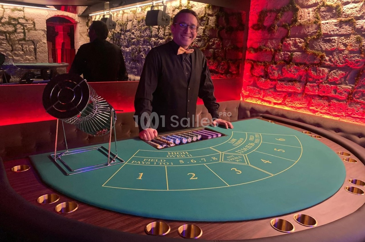 Homme souriant debout derrière une table de jeu de casino avec un cylindre de tirage et des jetons alignés.