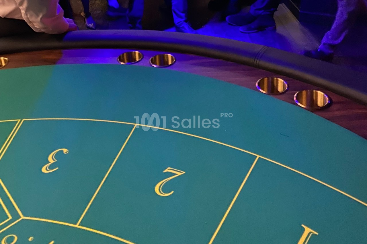 Table de jeu de casino avec tapis vert numéroté et porte-gobelets intégrés, entourée de personnes debout.
