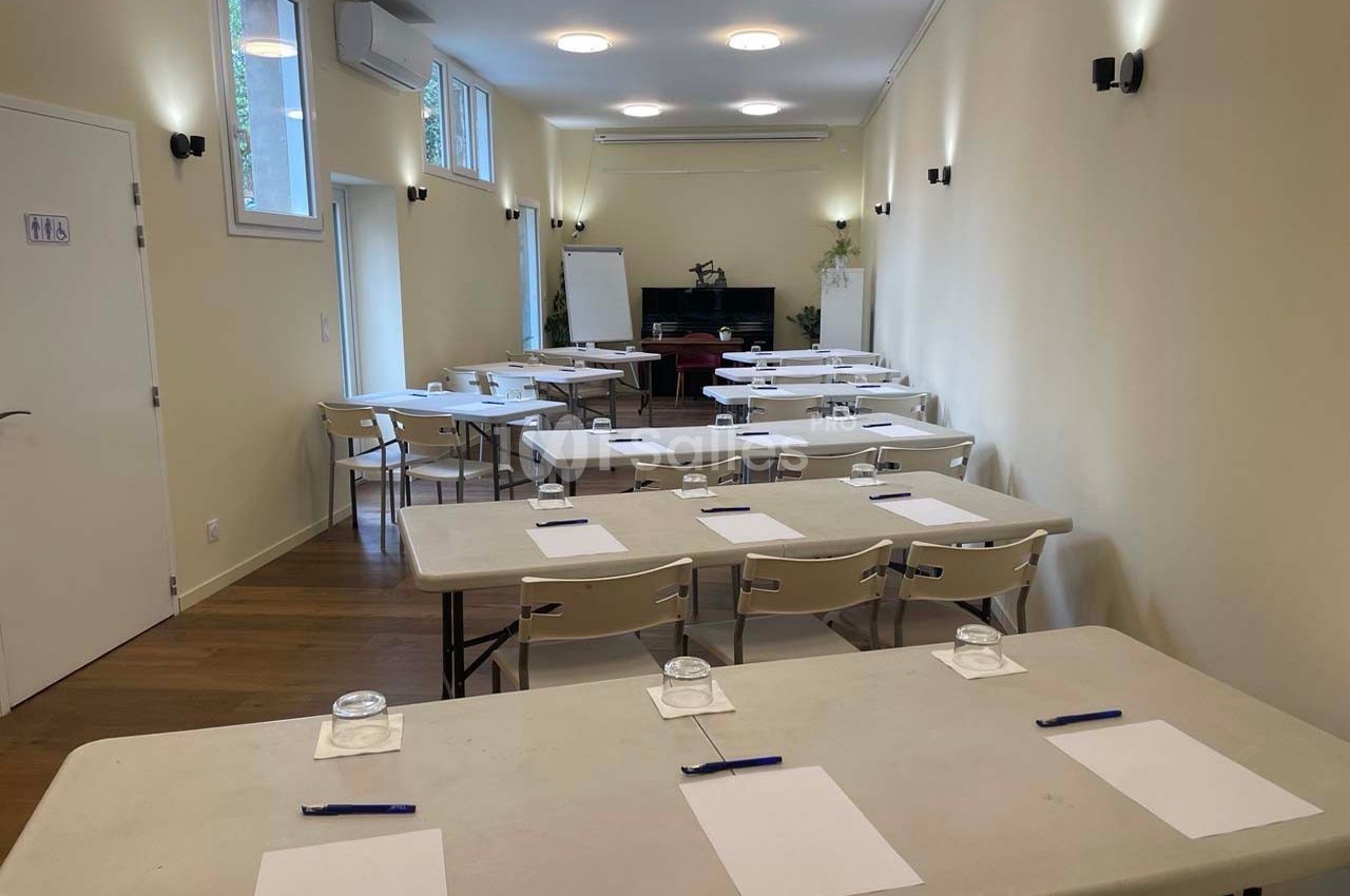 Salle lumineuse aménagée avec des tables, chaises, blocs-notes, stylos et verres, prête pour une réunion ou un atelier.