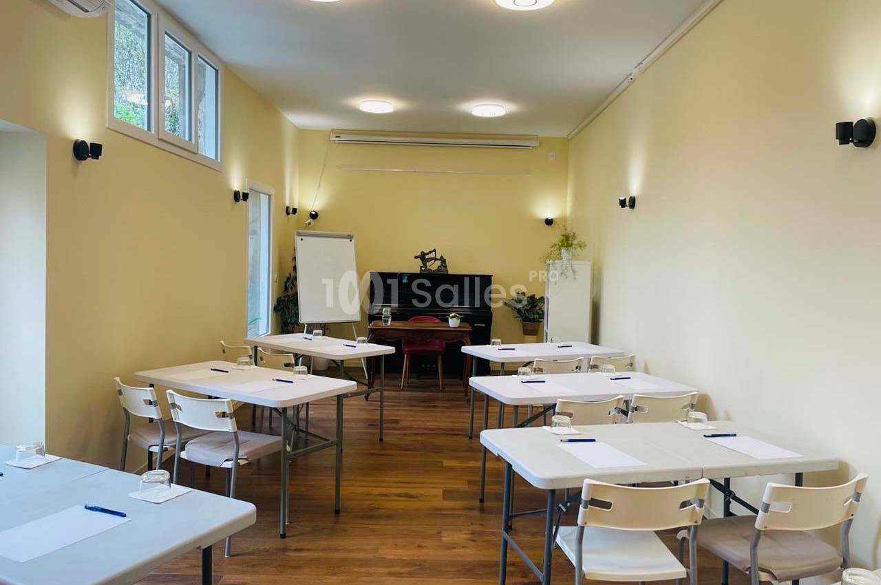 Salle lumineuse avec tables et chaises disposées pour une réunion ou une formation, tableau blanc et plantes en arrière-plan.