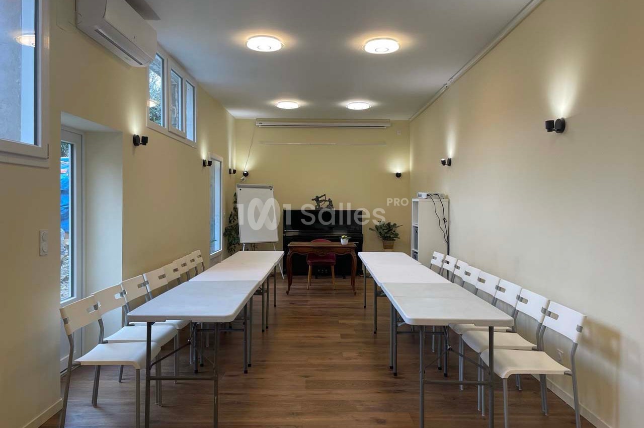 Salle lumineuse avec tables blanches disposées en U, chaises alignées, tableau blanc et piano en arrière-plan.