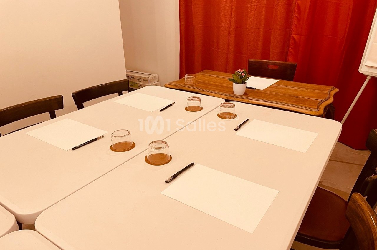 Table blanche avec des feuilles, stylos et verres disposés pour une réunion ou un atelier, devant un rideau rouge.